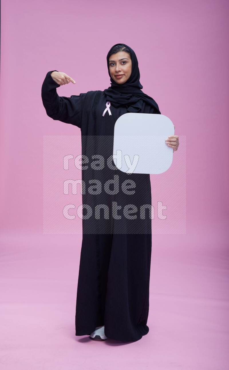 Saudi woman wearing pink ribbon on Abaya standing holding social media signs on pink background