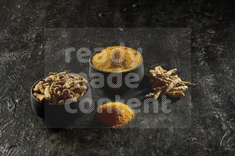 2 pottery black bowls, one full of turmeric powder and the other full of dried turmeric whole fingers on a textured black flooring