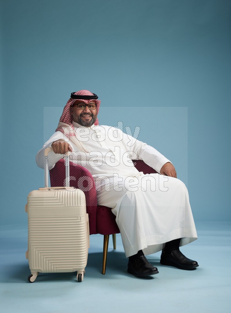 Saudi Man with shimag sitting on chair with a travel luggage on blue background