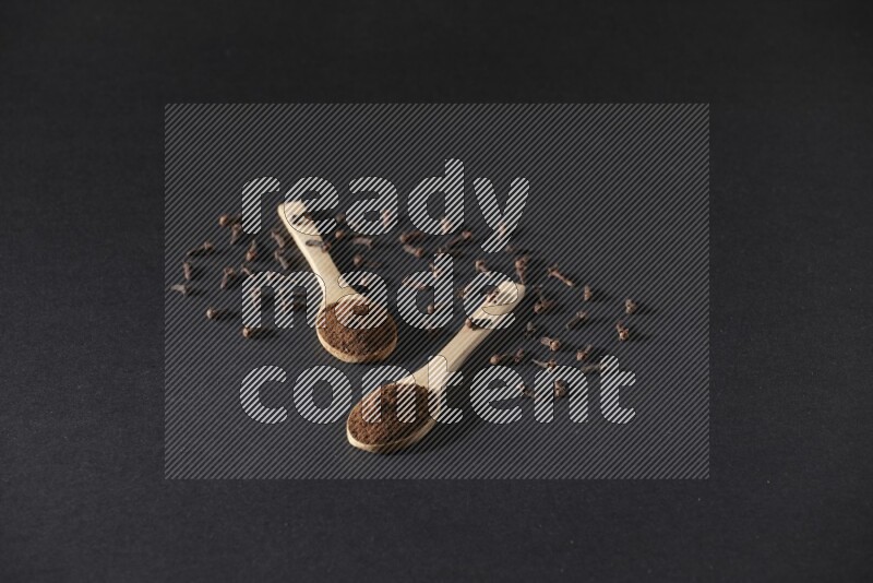 2 wooden spoons full of cloves powder with spreaded whole cloves on a black flooring