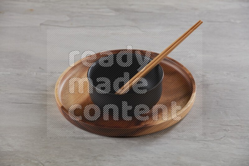 black ceramic round bowl on top of brown wood round plate and wood chopsticks, on grey textured countertop