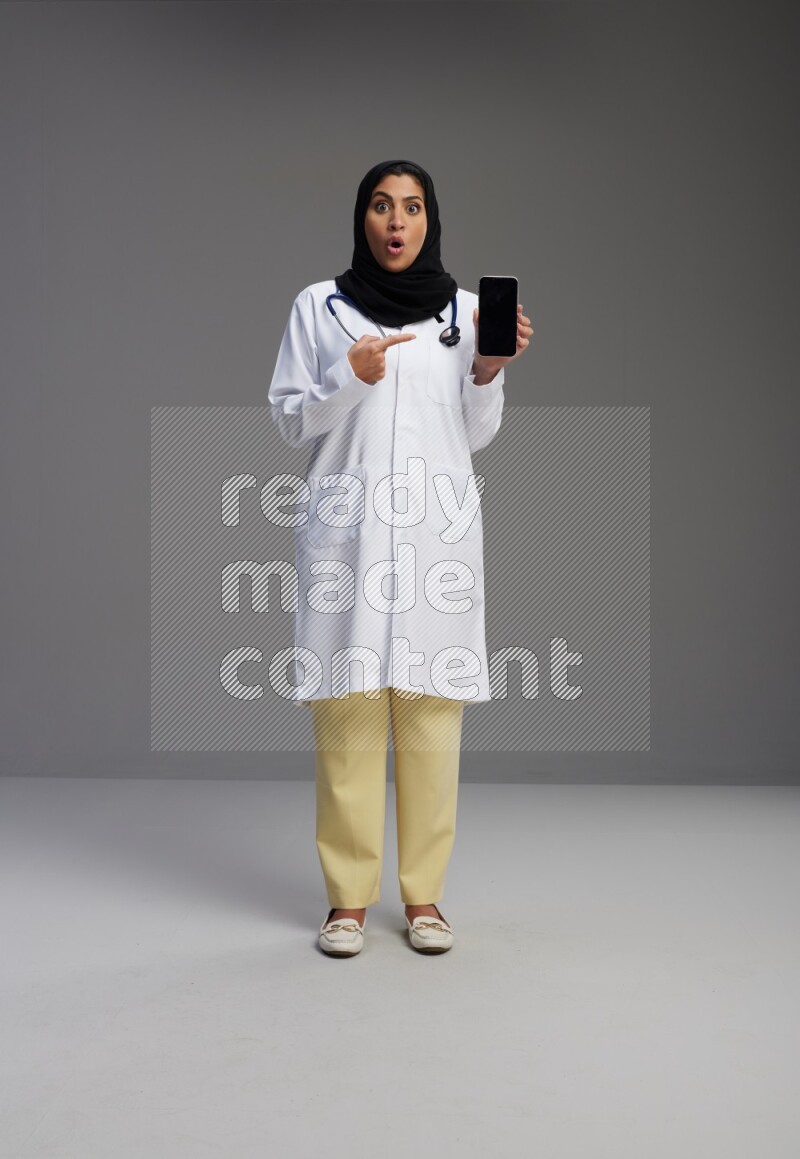 Saudi woman wearing lab coat with stethoscope standing showing phone to camera with sign in the back on Gray background