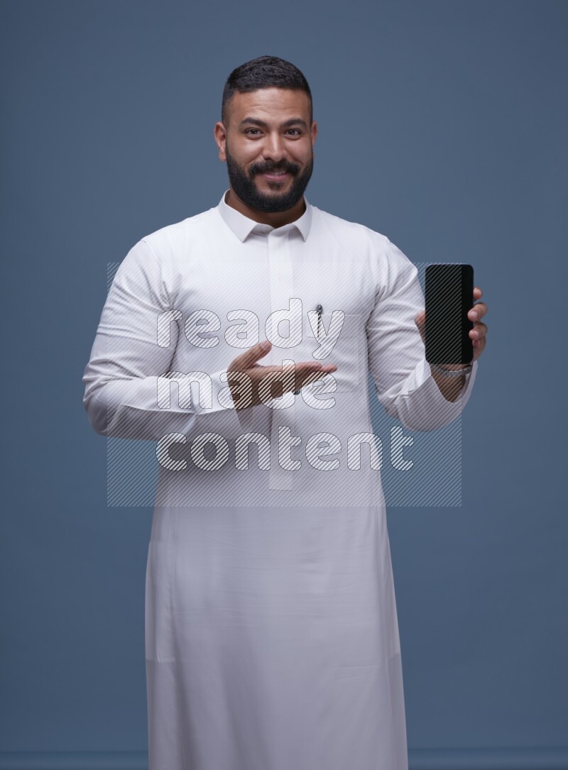A man Showing his phone in Blue Background wearing Saudi Thob