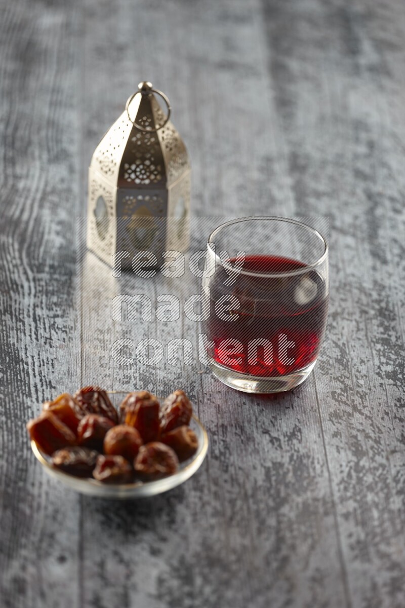 A silver lantern with different drinks, dates, nuts, prayer beads and quran on grey wooden background