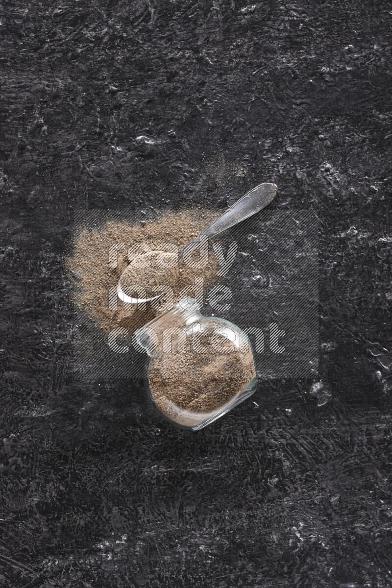 A flipped glass spice jar full of black pepper powder with a metal spoon full of powder on textured black flooring