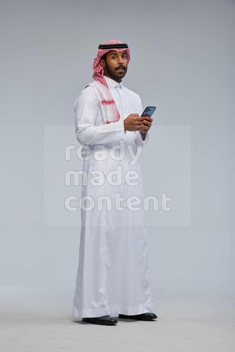 Saudi man Wearing Thob and shomag standing texting on phone on Gray background