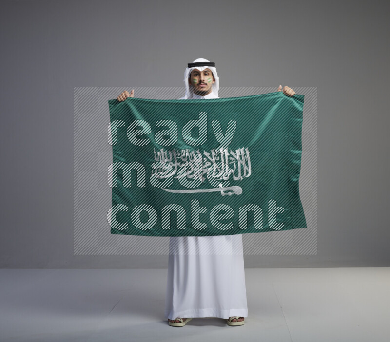 A Saudi man standing wearing thob and white shomag with face painting holding big Saudi flag on gray background