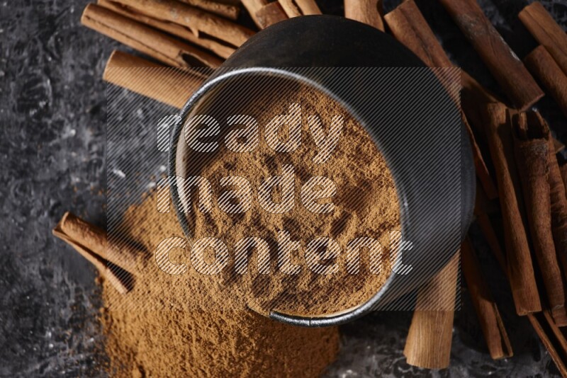 Black pottery bowl over filled with cinnamon powder and cinnamon sticks around the bowl on a textured black background