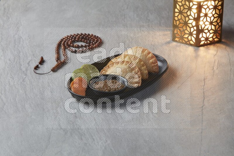 Three fried sambosas in an oval shaped black plate and a red sauce in a black round ramekin with a brown misbaha and a golden lantern on the side on a gray background