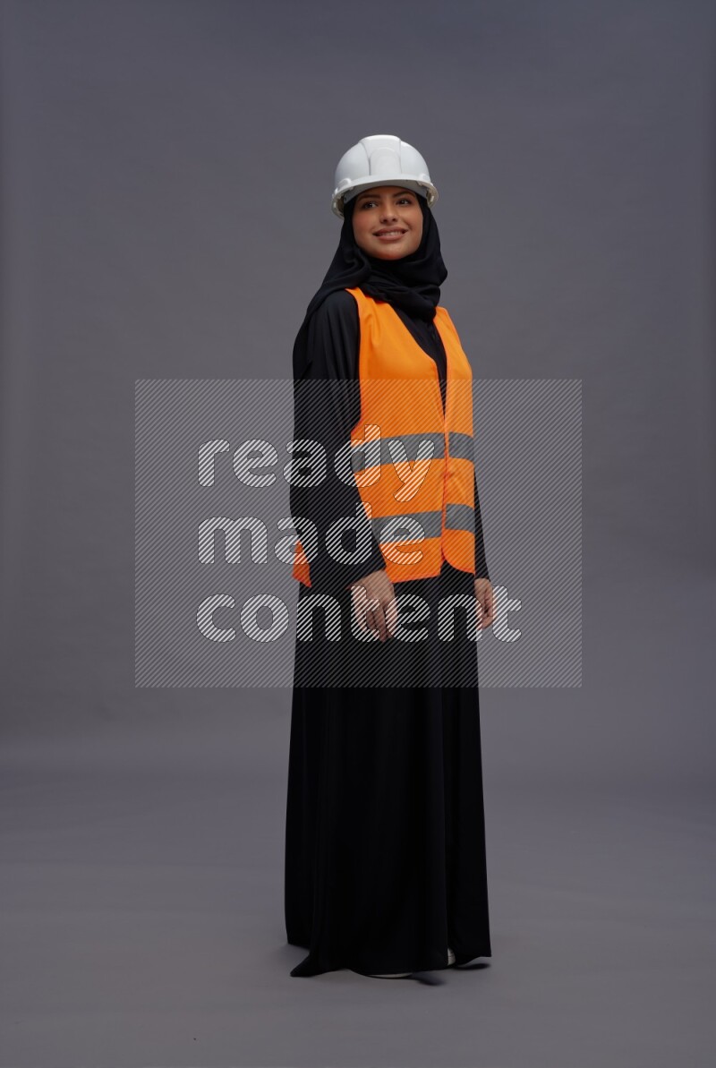 Saudi woman wearing Abaya with engineer vest standing interacting with the camera on gray background