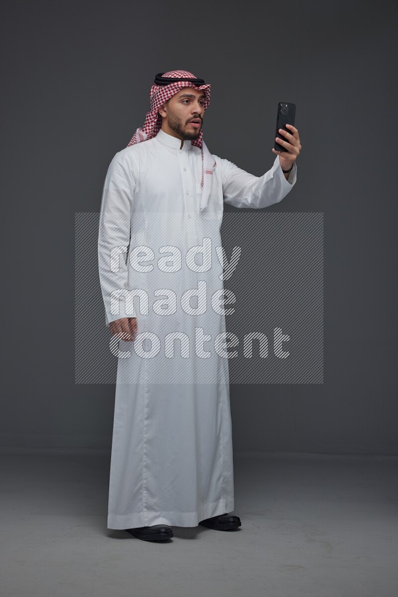 A Saudi man wearing Thobe and Shamgh taking selfie with his phone eye level on a gray background