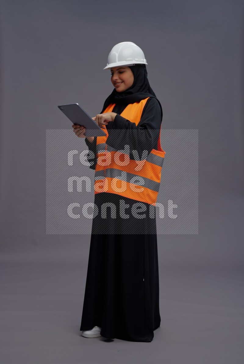 Saudi woman wearing Abaya with engineer vest standing working on tablet on gray background