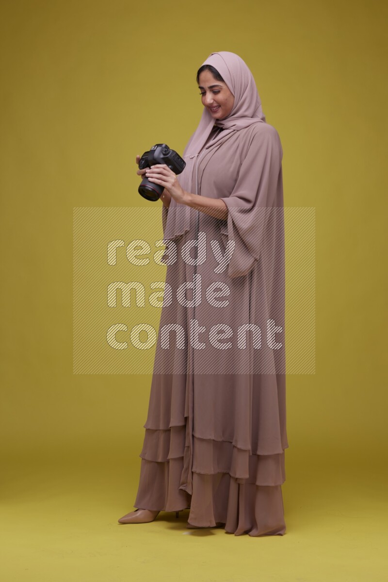 A woman Holding a Camera on a Yellow Background wearing Brown Abaya with Hijab