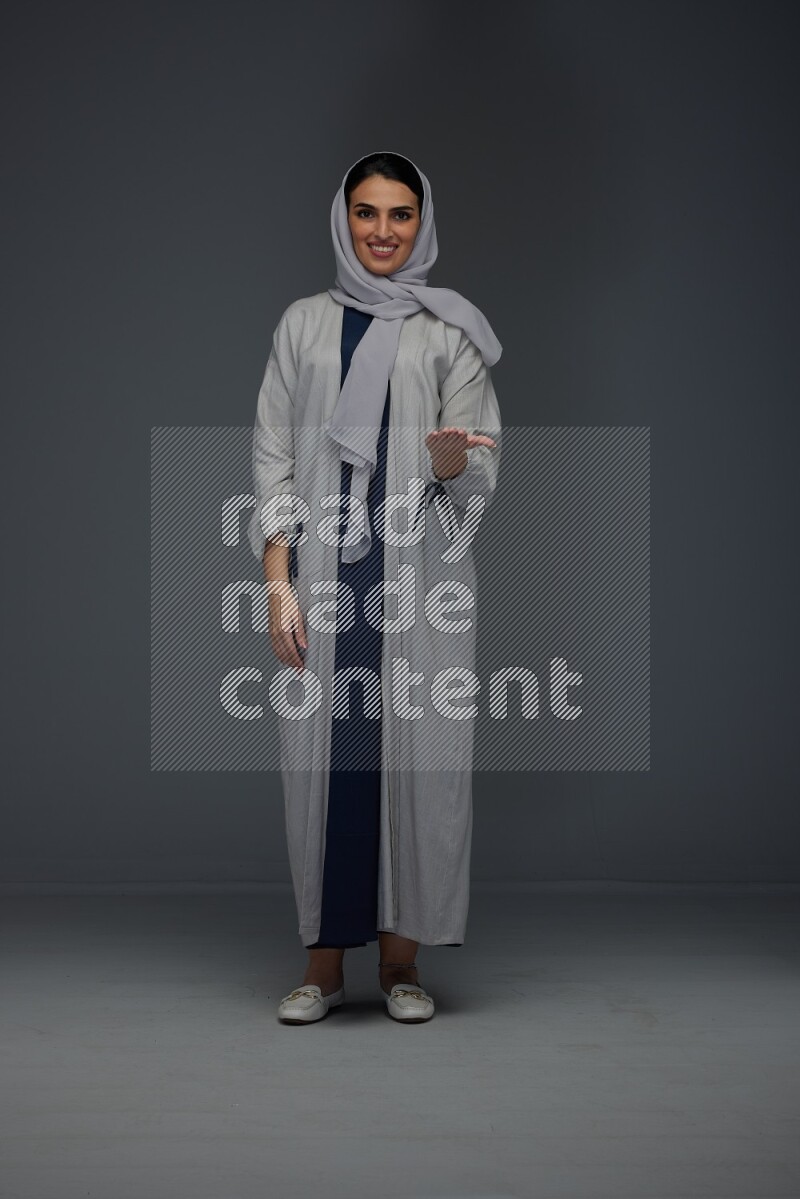A Saudi woman wearing a light gray Abaya and head scarf standing and pointing in different directions eye level on a grey background
