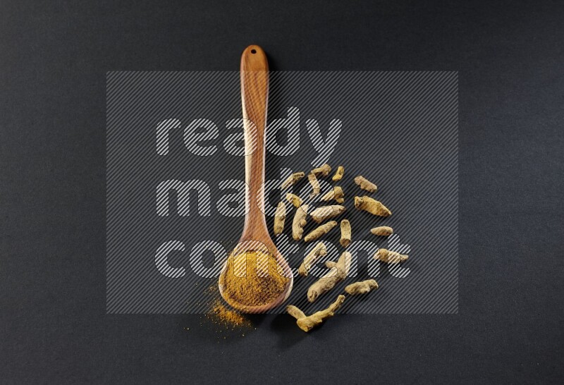 A wooden ladle full of turmeric powder with dried turmeric fingers on black flooring