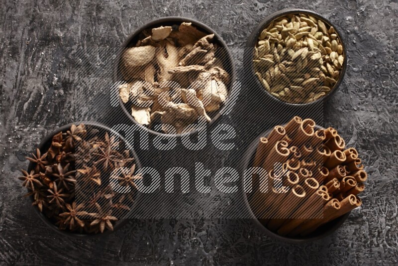 Ginger, Cardamom, Star anise and cinnamon sticks in 4 bowls on a textured black background