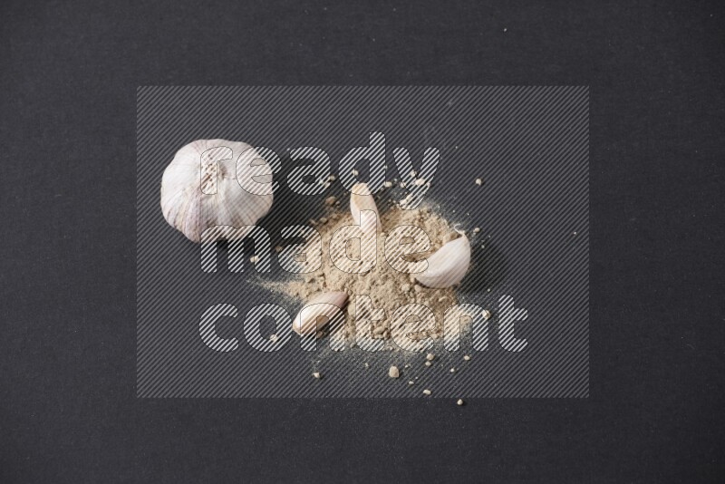 A small pile of garlic powder with some of cloves and a whole garlic bulb on a black flooring