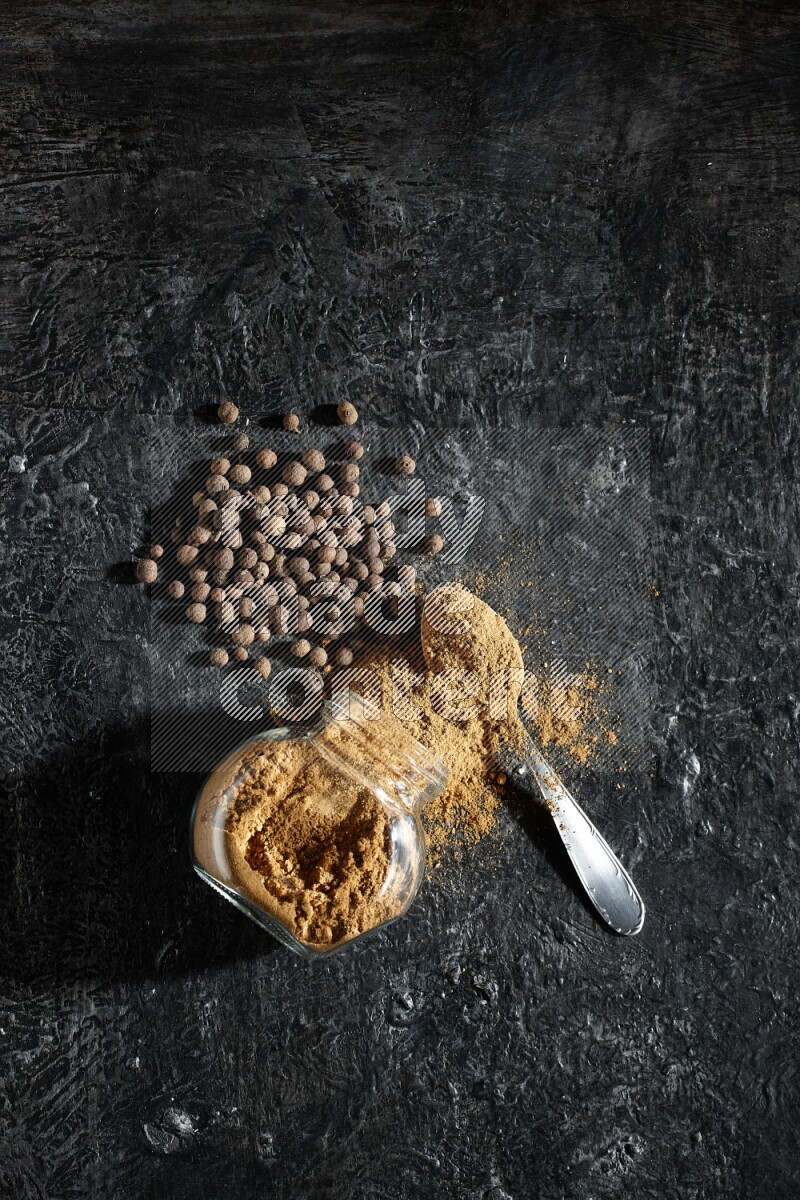 A flipped glass spice jar and metal spoon full of allspice powder and powder spilled out of it with whole balls on a textured black flooring