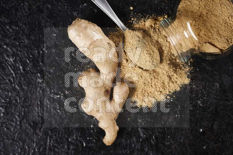 A glass jar full of ground ginger powder flipped with some spilling powder on black background