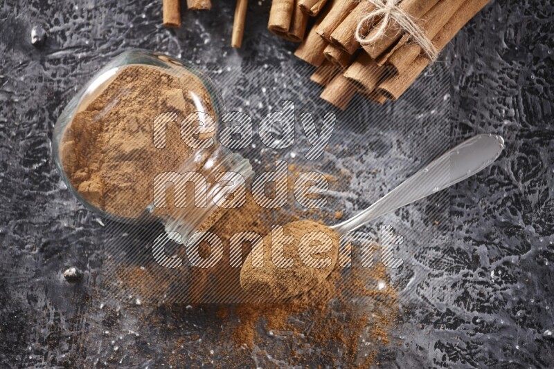 Herbal glass jar full cinnamon powder flipped and a metal spoon full of powder, cinnamon sticks stacked and bounded in the back on textured black background in different angles