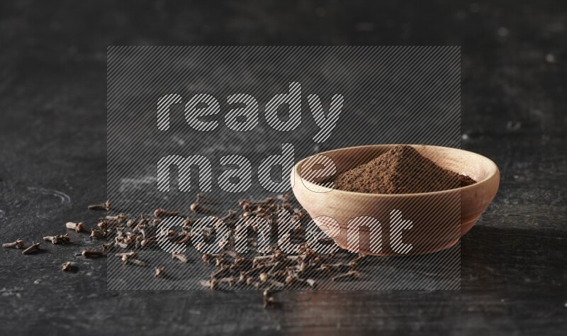 A wooden bowl full of cloves powder on a textured black flooring
