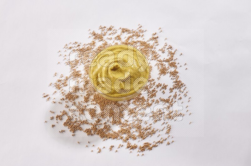 A glass bowl full of mustard paste with mustard seeds underneath on white flooring