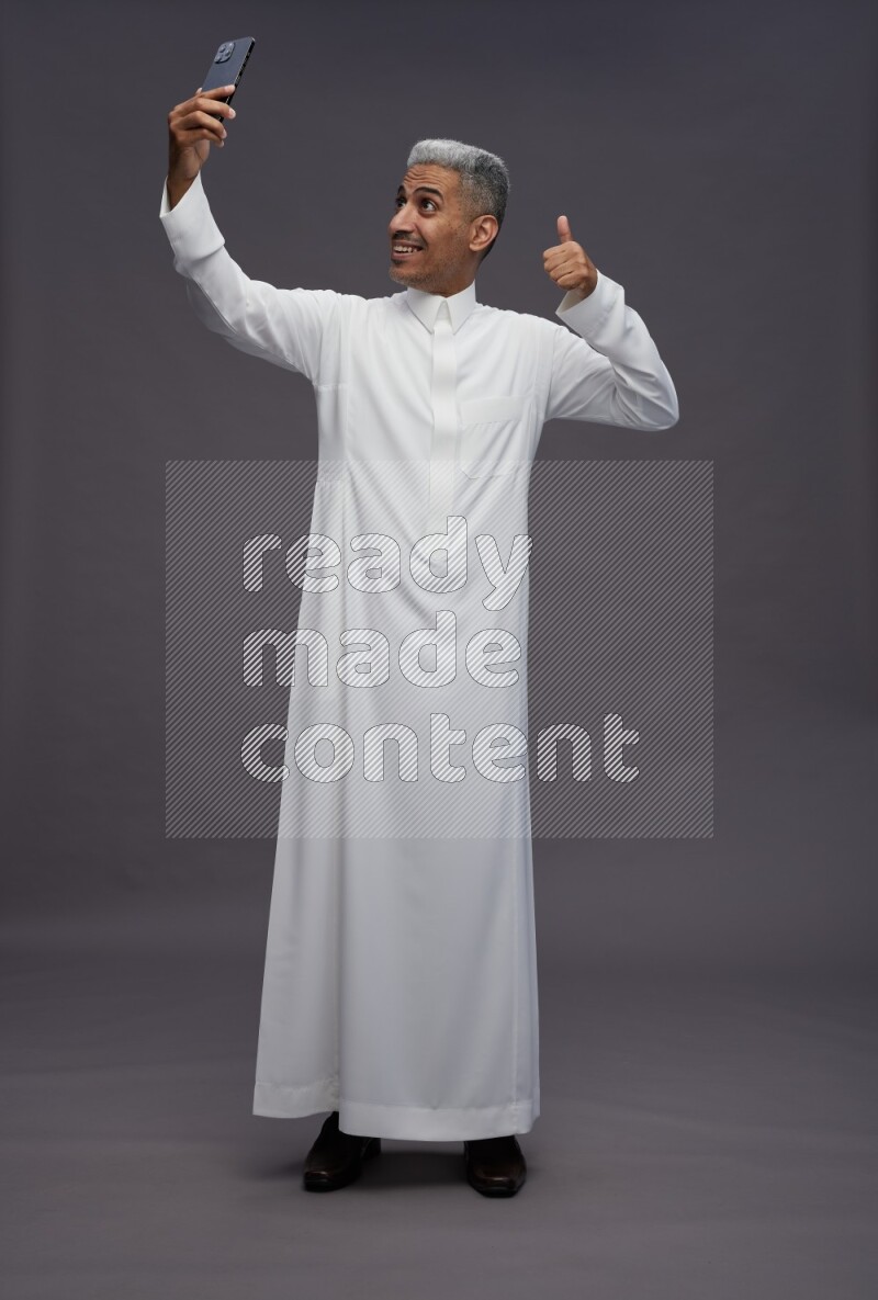 Saudi man wearing thob standing taking selfie on gray background