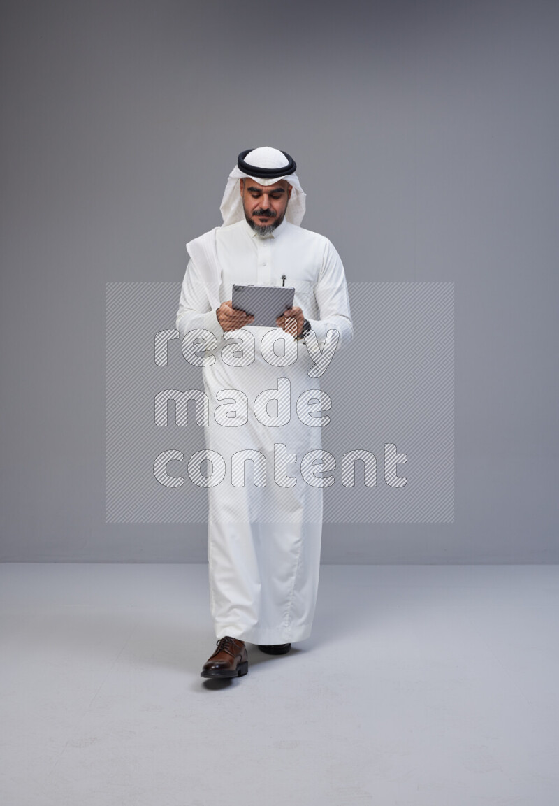 Saudi man Wearing Thob and white Shomag standing working on tablet on Gray background