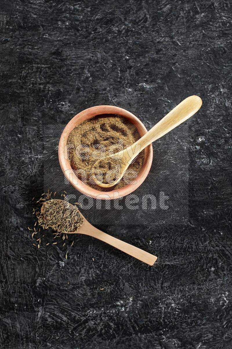 A wooden bowl and 2 wooden spoons full of cumin powder and cumin seeds on a textured black flooring