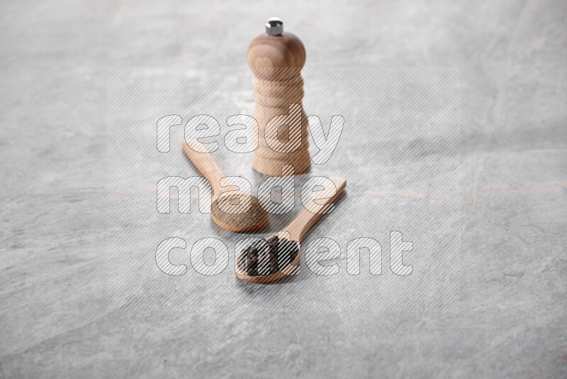 2 wooden spoons, one full of black pepper powder and the other full of black pepper beads and wooden grinder on a marble flooring