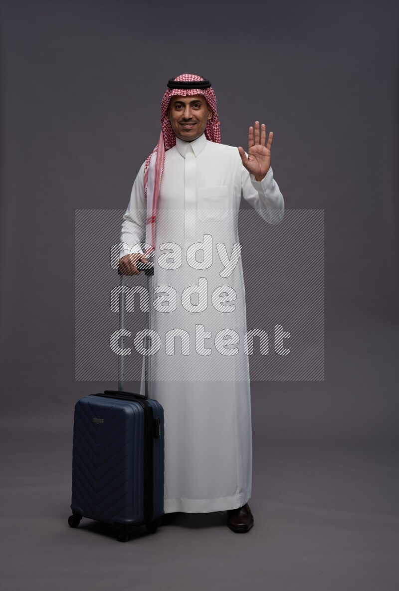 Saudi man wearing thob and shomag standing holding bag on gray background
