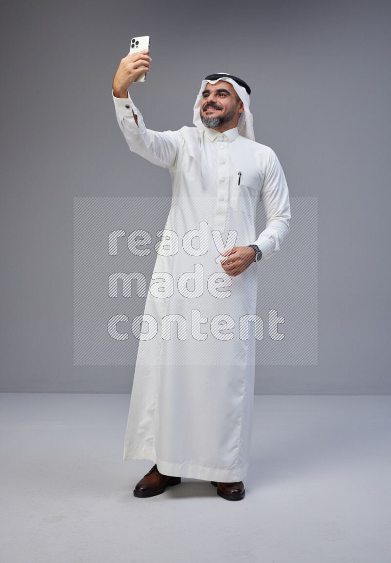 Saudi man Wearing Thob and white Shomag standing taking selfie on Gray background
