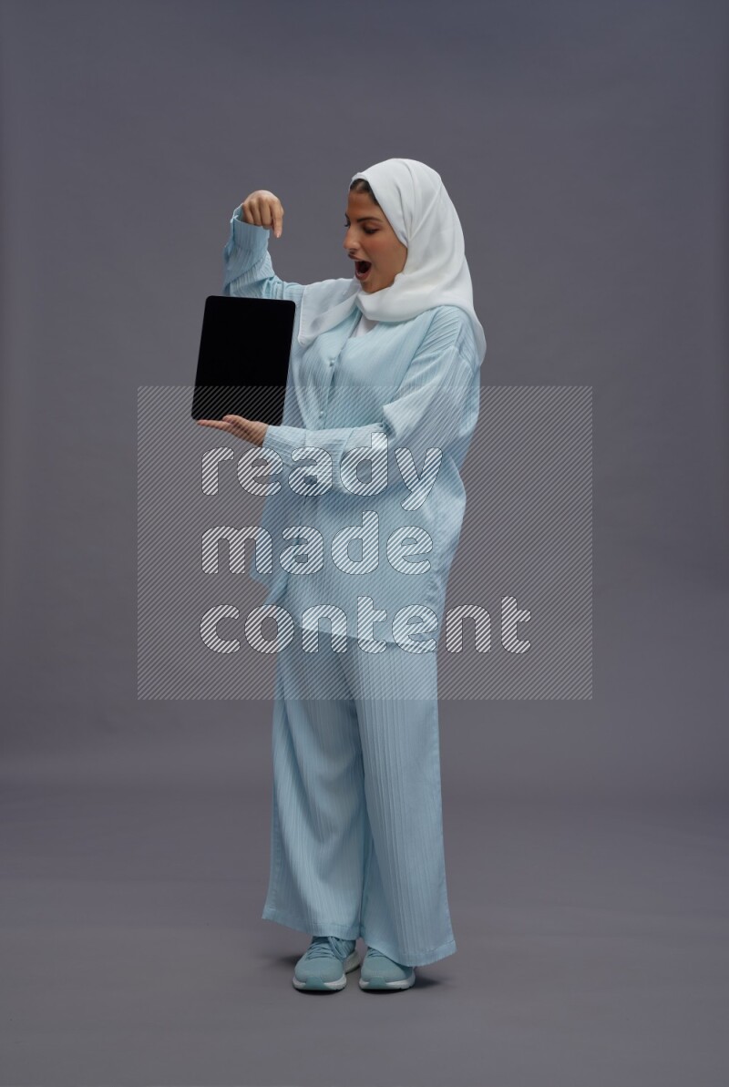 Saudi woman wearing hijab clothes standing showing tablet to camera on gray background