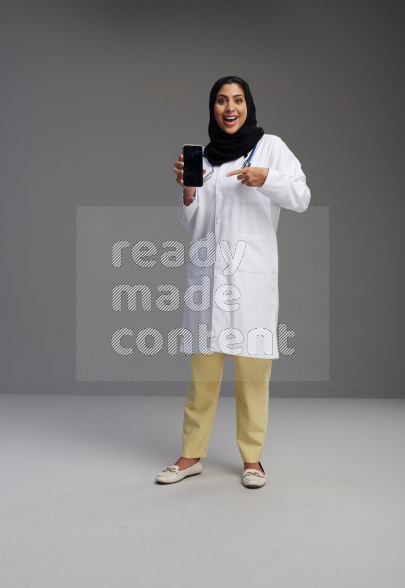 Saudi woman wearing lab coat with stethoscope standing showing phone to camera with sign in the back on Gray background