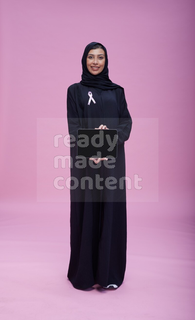 Saudi woman wearing pink ribbon on Abaya standing showing a tablet to camera on pink background