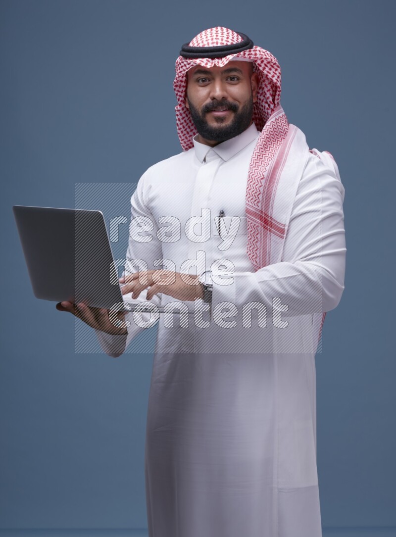 A man standing  with a laptop on Blue Background wearing Saudi Thob and Shomag