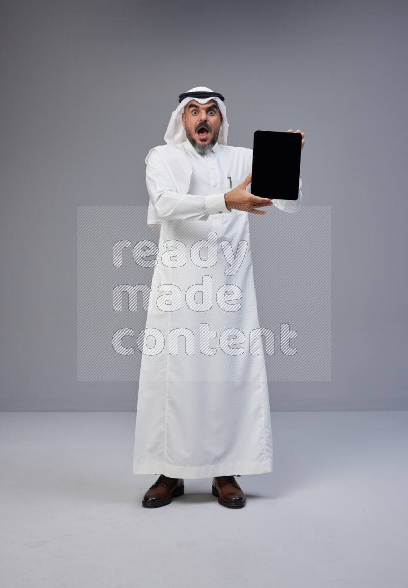 Saudi man Wearing Thob and white Shomag standing showing tablet to camera on Gray background