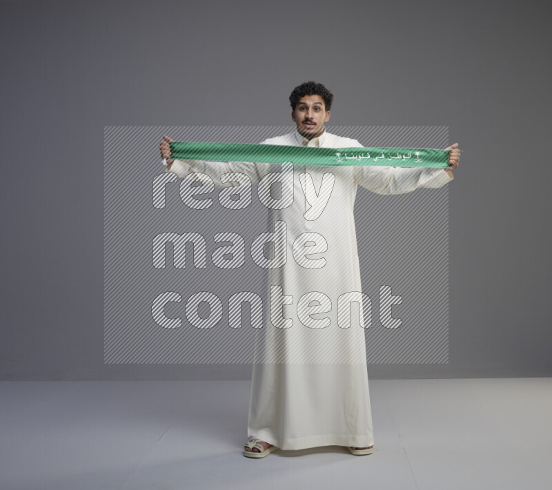 A Saudi man standing wearing thob holding Saudi flag scarf on gray background
