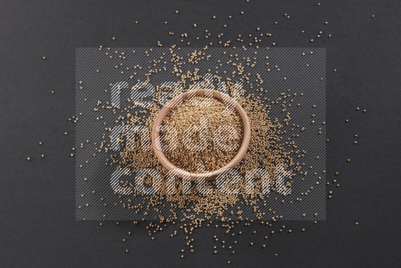 A wooden bowl full of mustard seeds and more seeds spread on a black flooring