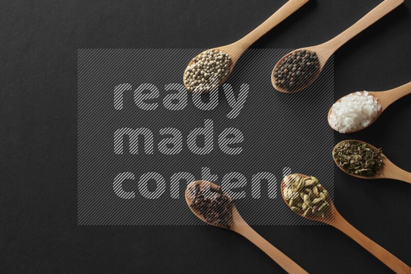 wooden spoons filled with white peppers, cloves, cardamom, salt, black peppers and basil on black flooring