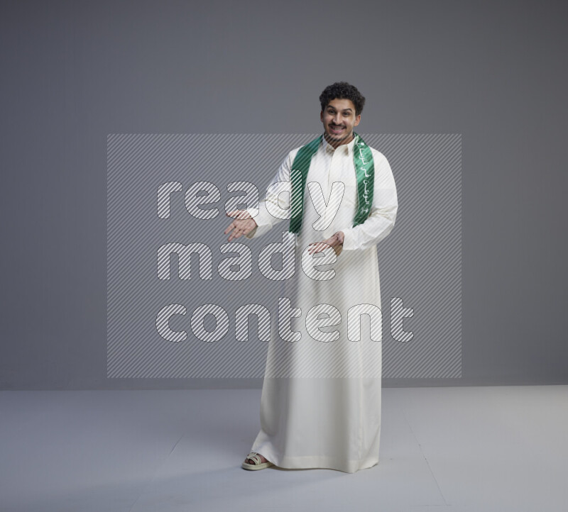 A Saudi man standing wearing thob and Saudi flag scarf on gray background