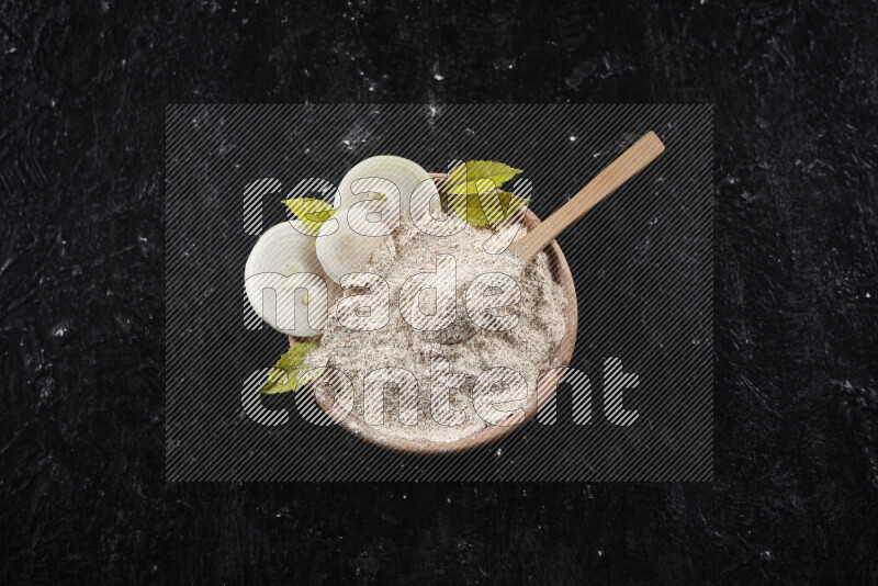 A wooden bowl full of onion powder with a wooden spoon in it with some sliced onions on black background