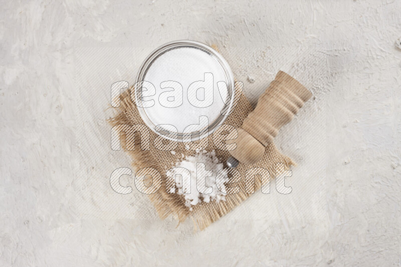 A glass bowl full of white salt with a wooden grinder on a burlap fabric all on white background