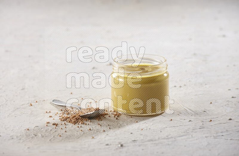 A glass jar full of mustard paste and a metal spoon full of mustard seeds on a textured white flooring
