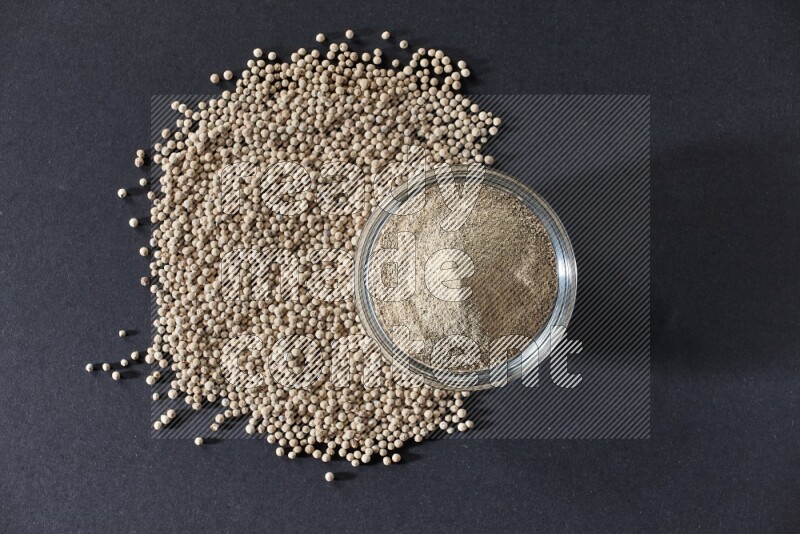 A glass bowl full of white pepper powder with white pepper beads on black flooring