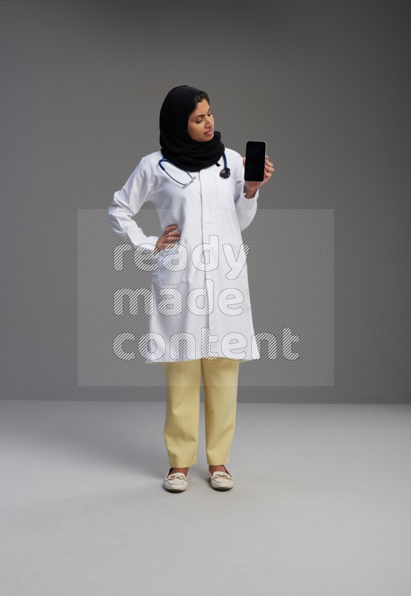 Saudi woman wearing lab coat with stethoscope standing showing phone to camera with sign in the back on Gray background