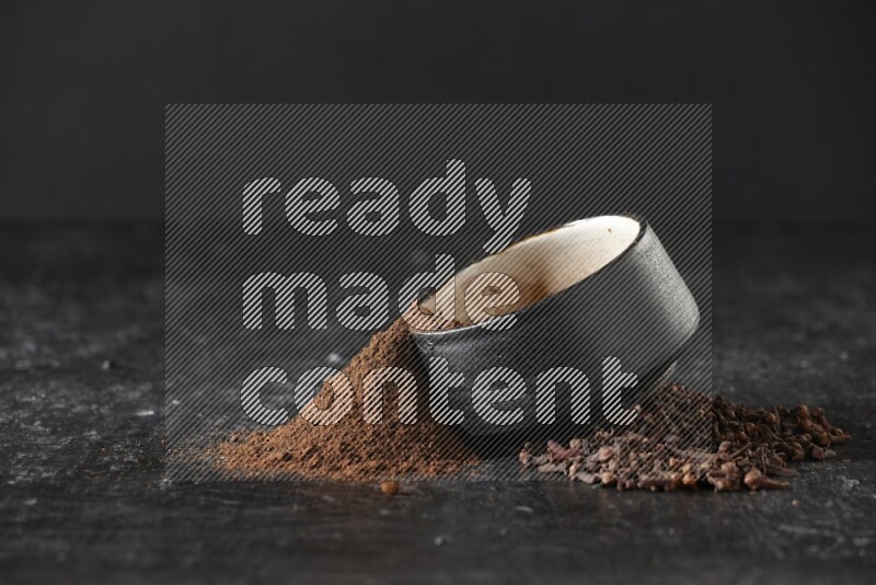 A black pottery bowl full of cloves powder spilled out of it with whole grains on a textured black flooring