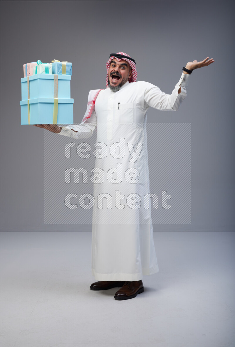 Saudi man Wearing Thob and red Shomag standing holding gift box on Gray background