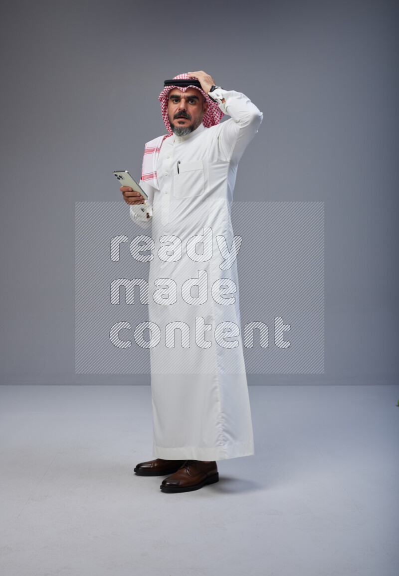 Saudi man Wearing Thob and red Shomag standing texting on phone on Gray background