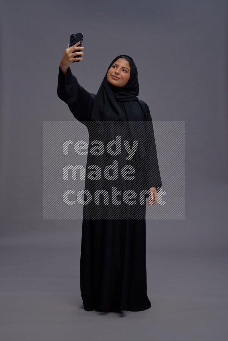 Saudi woman wearing Abaya standing taking selfie on gray background